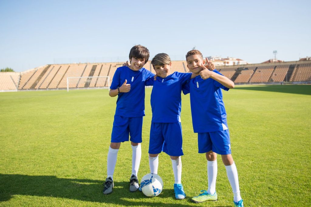 Three young boys in blue uniforms posing with a soccer ball on a sunny day.